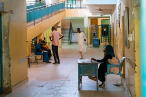 Bashair Hospital hallway with a nurse seated on a blue chair with an open book on the table at the fore ground with patients and nurses interacting in the far ground.