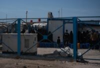 A blue mesh fence in front of people queuing with jerrycans in Al-Hol Camp, Eastern Al-Hasakah Governorate, Northeastern Syria.