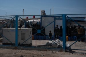 A blue mesh fence in front of people queuing with jerrycans in Al-Hol Camp, Eastern Al-Hasakah Governorate, Northeastern Syria.