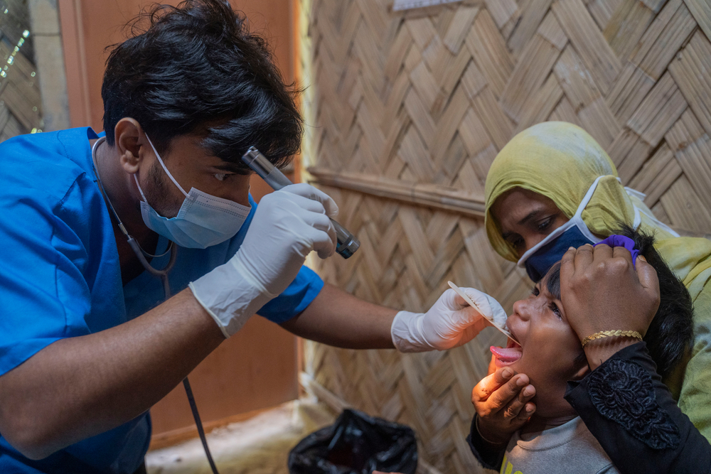 A medical professional wearing scrubs, a medical mask and gloves performs a medical examination on a child as their mother helps.