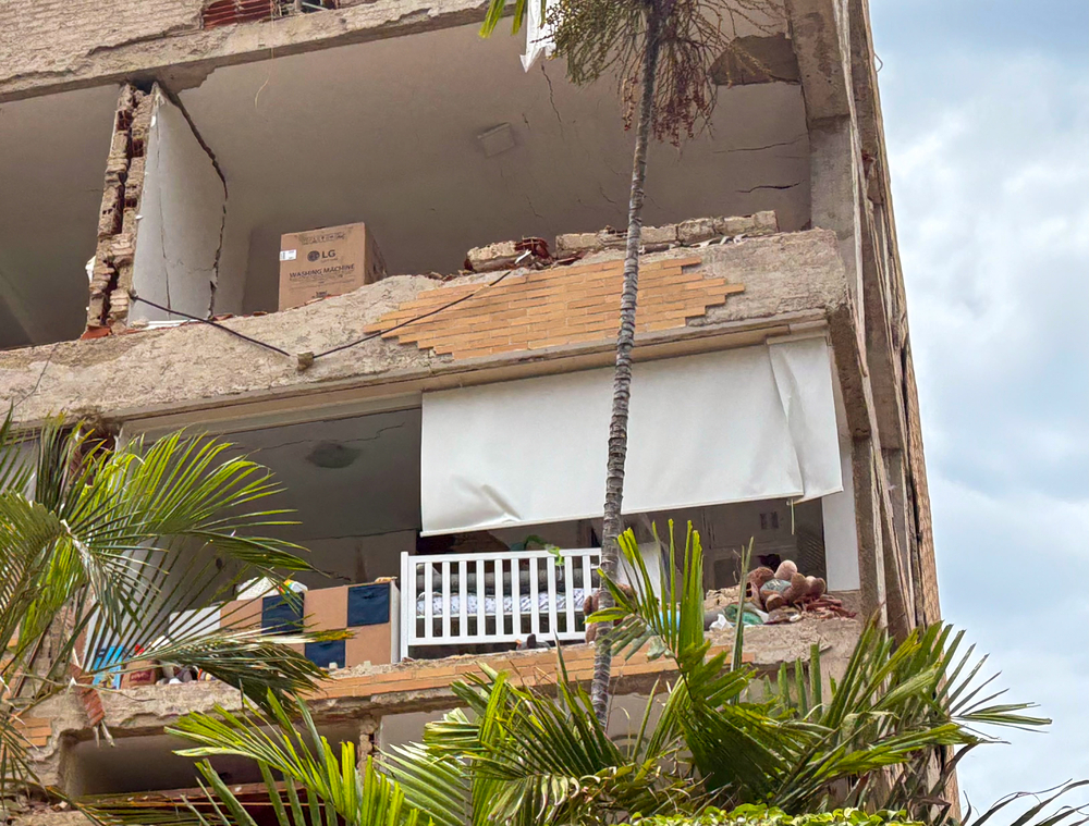 A damaged building with a collapsed wall exposes a room containing a baby’s cot. Venezuela, 2026.